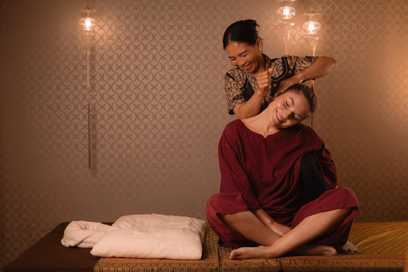 A Thai massage therapist eases the shoulders of a seated, clothed client