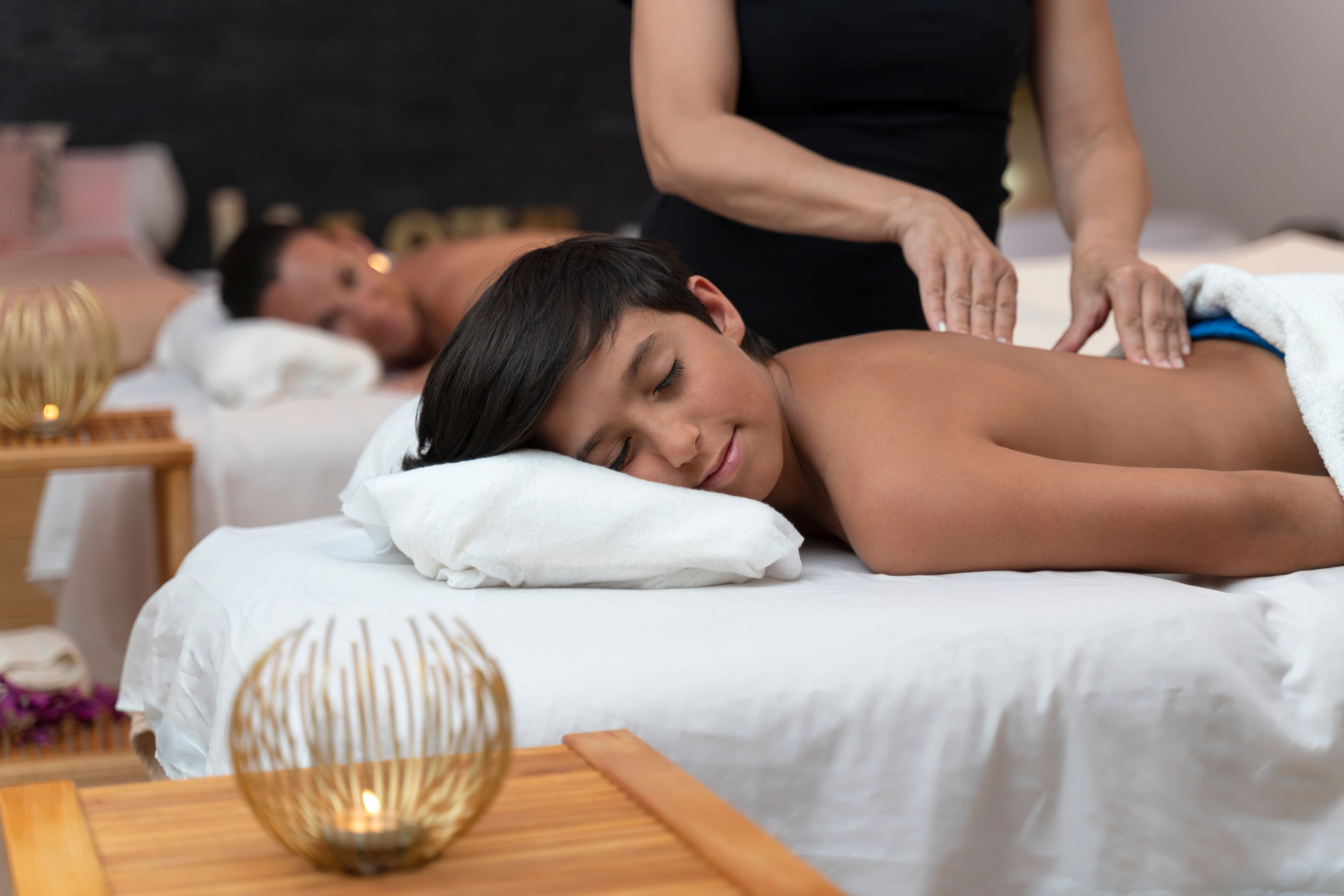 A young guest has a gentle spa massage while a parent relaxes nearby