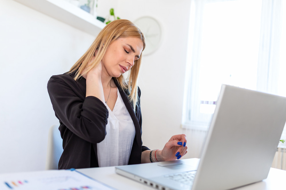An office worker rubbing her stiff neck at her desk, a sign she needs a lunch break massage.